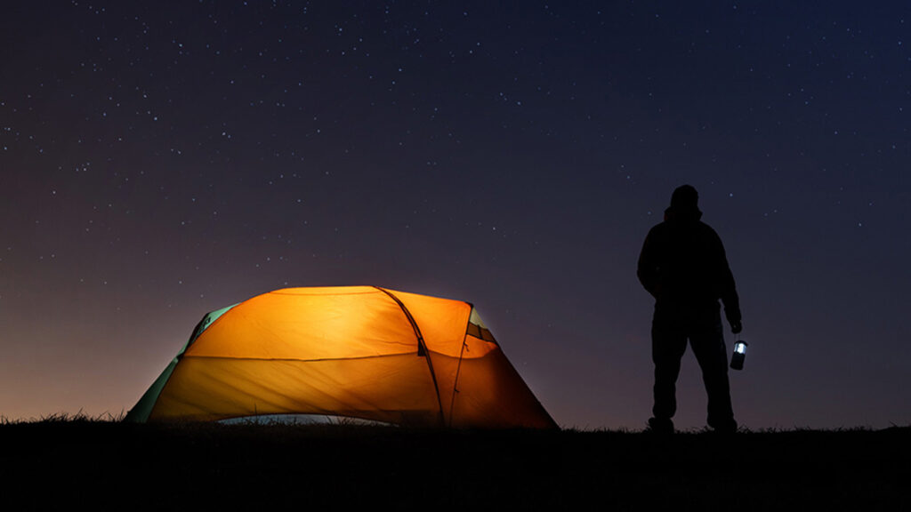 A silhouette of a man standing next to a tent on a starry night.
